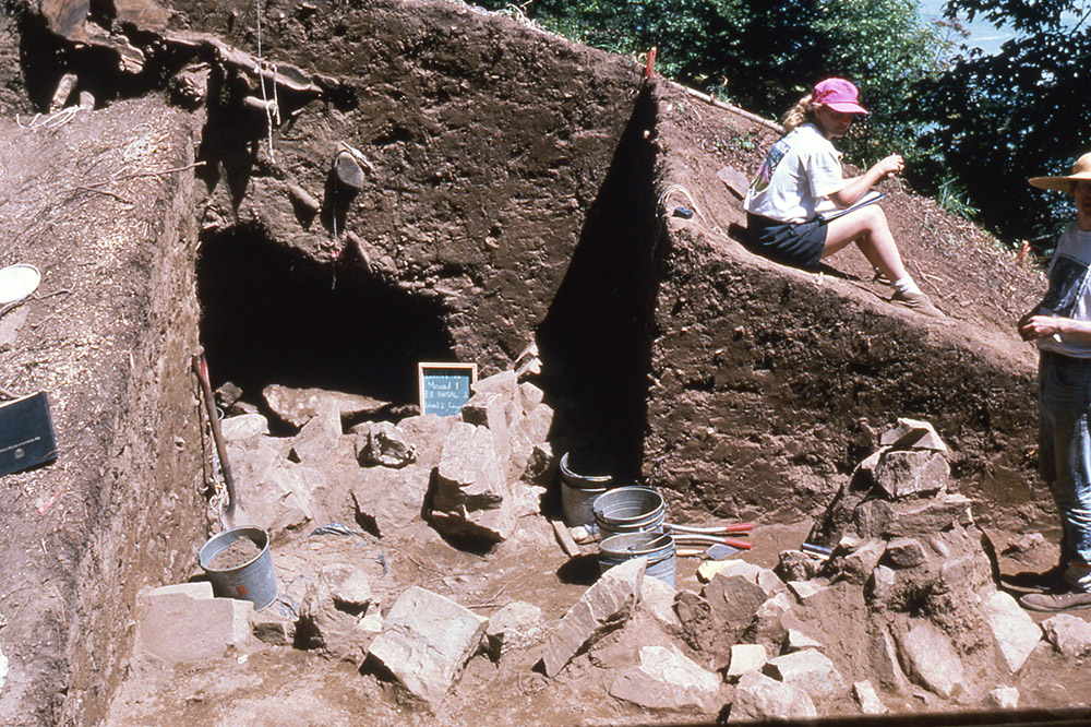 Une femme assise à côté d’une section de terre qui a été creusée. À l’intérieur de la section se trouve un gros tas de pierres.
