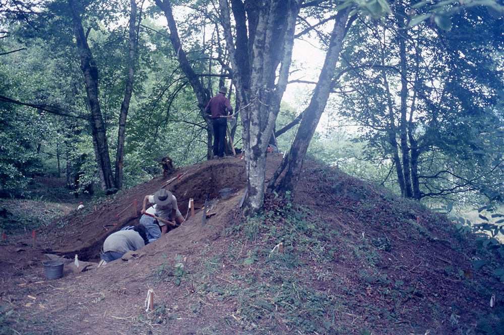 A group of archaeologists excavate a section of an earthen mound.