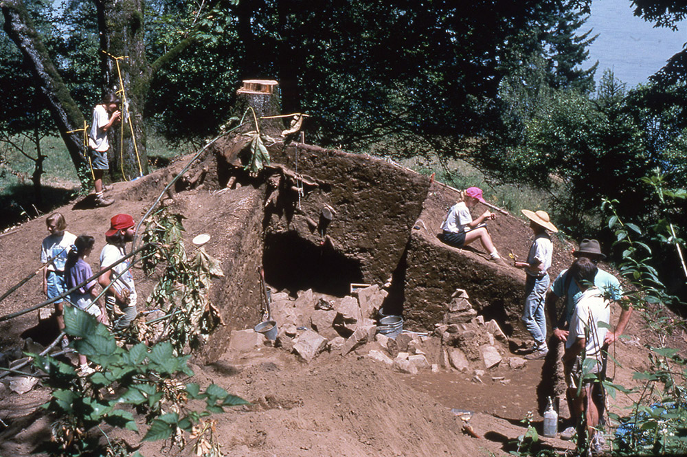 A group of archaeologists stand around the excavated sections of earth from a very large mound.