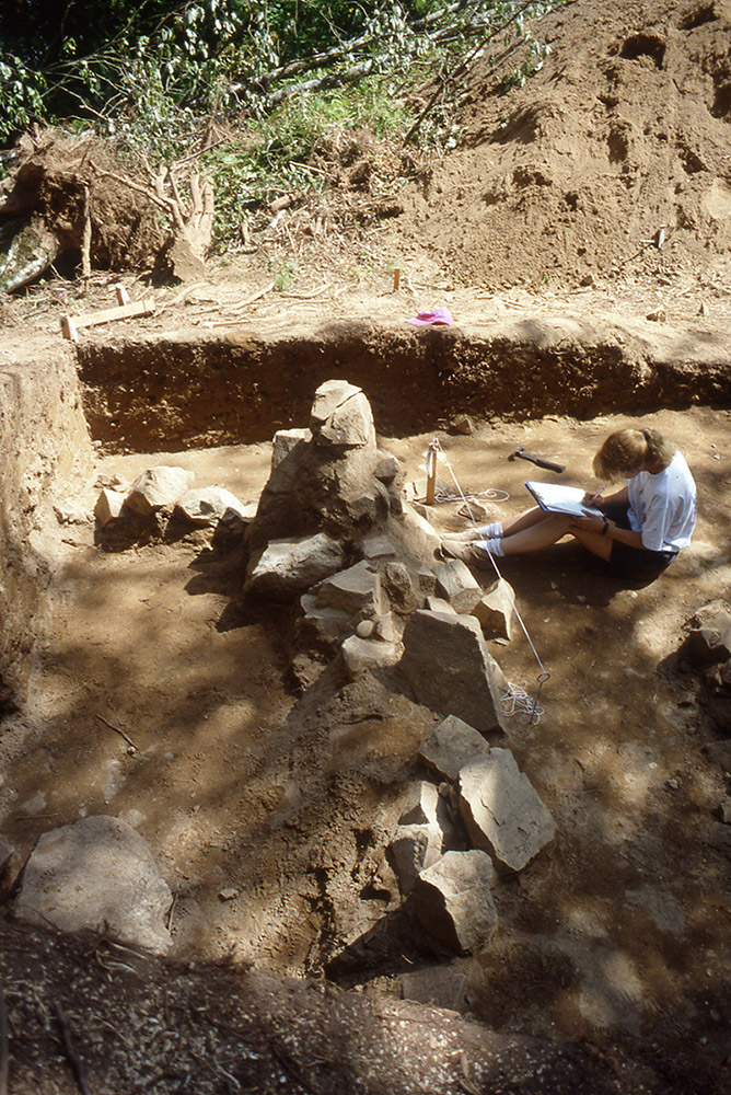 A woman takes field notes in front of a rock formation. She is seated beside an excavated mound. 
