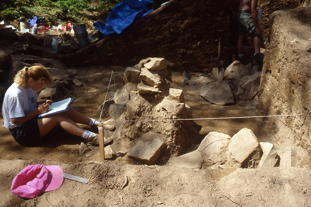 Une femme rédige des observations de terrain devant une formation rocheuse. Elle est assise devant une section creusée.