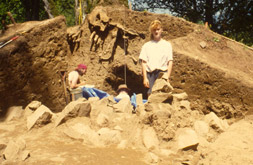 Several people are gathered alongside an excavated large mound of earth. The mound is 3 m tall and has been sectioned in half lengthwise. There are many large rocks towards the base of the mound.