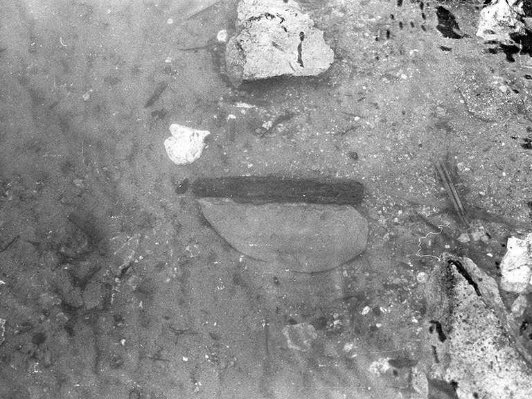 A black and white photograph shows an ancient slate knife with a wooden handle in the riverbank where it was found.