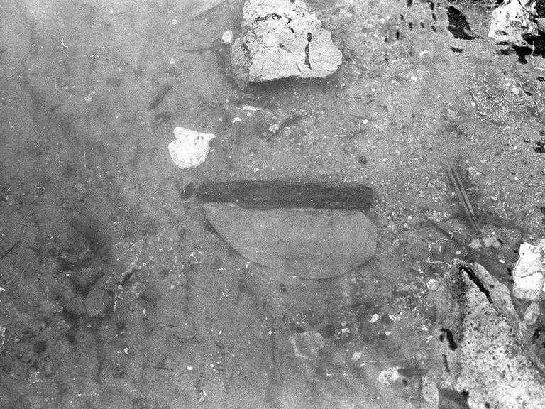 Une photo en noir et blanc montre un ancien couteau en pierre avec un manche en bois sur la berge où il a été trouvé.