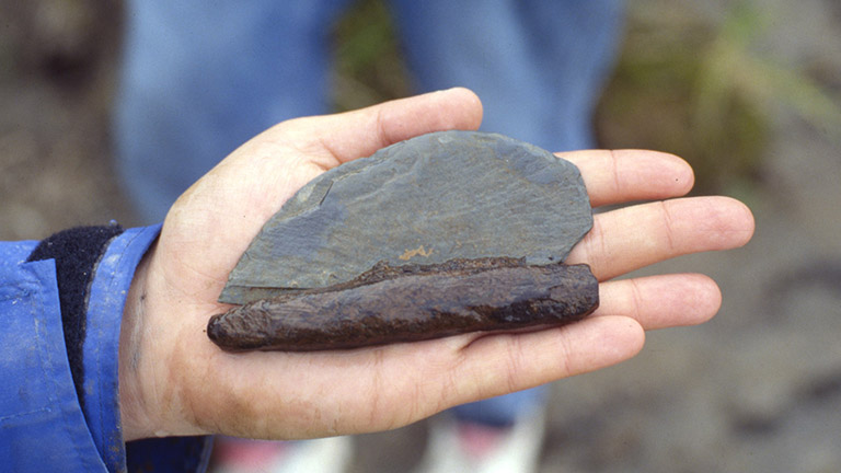 An ancient stone knife in its wooden handle is held in someone’s hand.