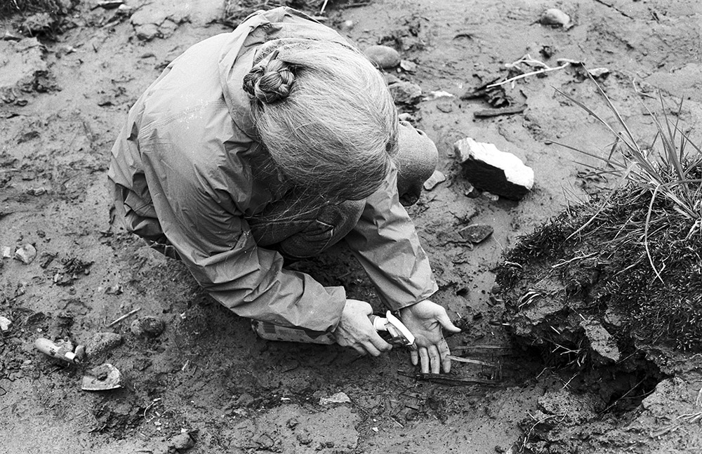 Une photo en noir et blanc d’une femme dans un secteur archéologique. Elle vaporise de l’eau sur les vestiges de vannerie pour les exposer.