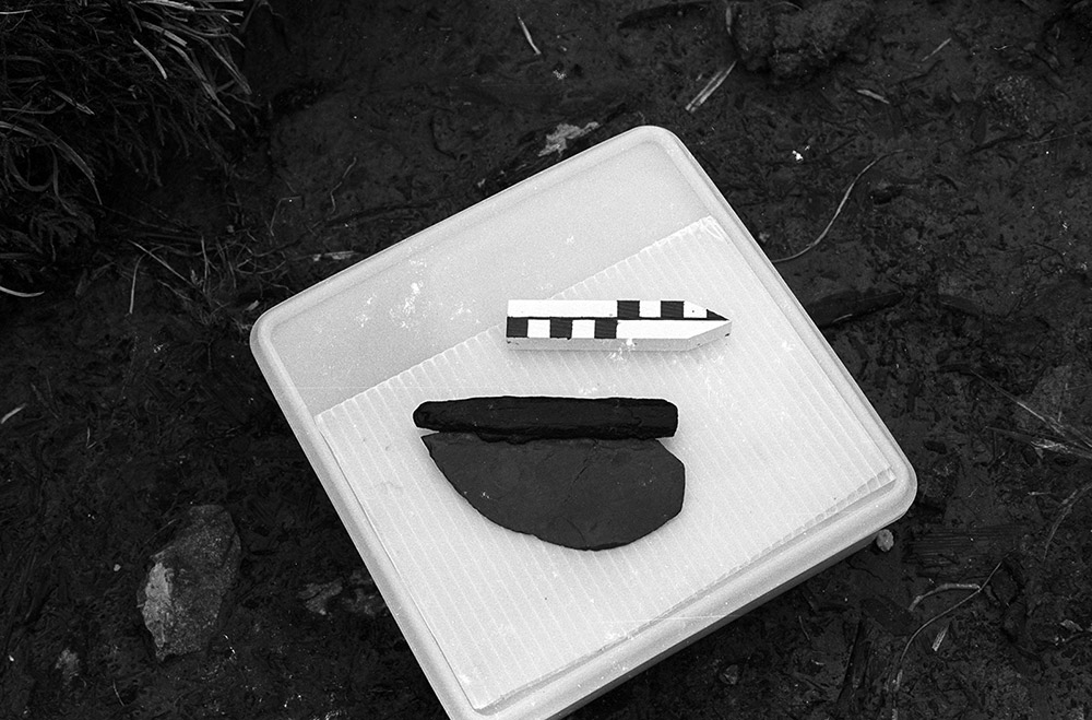 A black and white photograph shows an ancient stone knife with a wooden handle in the archaeological area it was found.