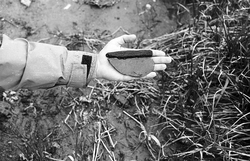 A black and white photograph shows someone holding an ancient stone knife with a wooden handle in the palm of their hand.
