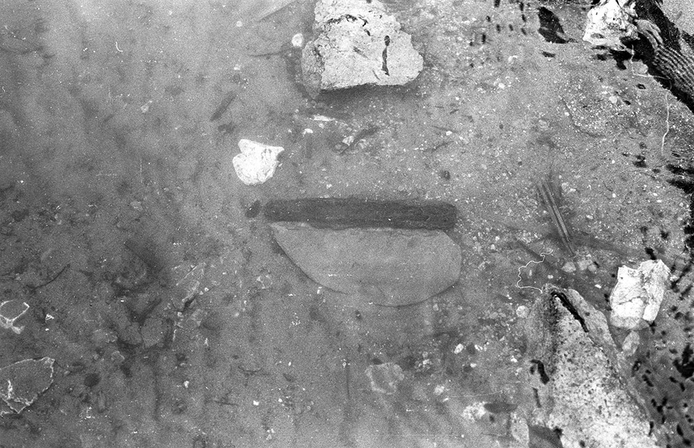 Une photo en noir et blanc montre un ancien couteau en pierre avec un manche en bois sur la berge où il a été trouvé.