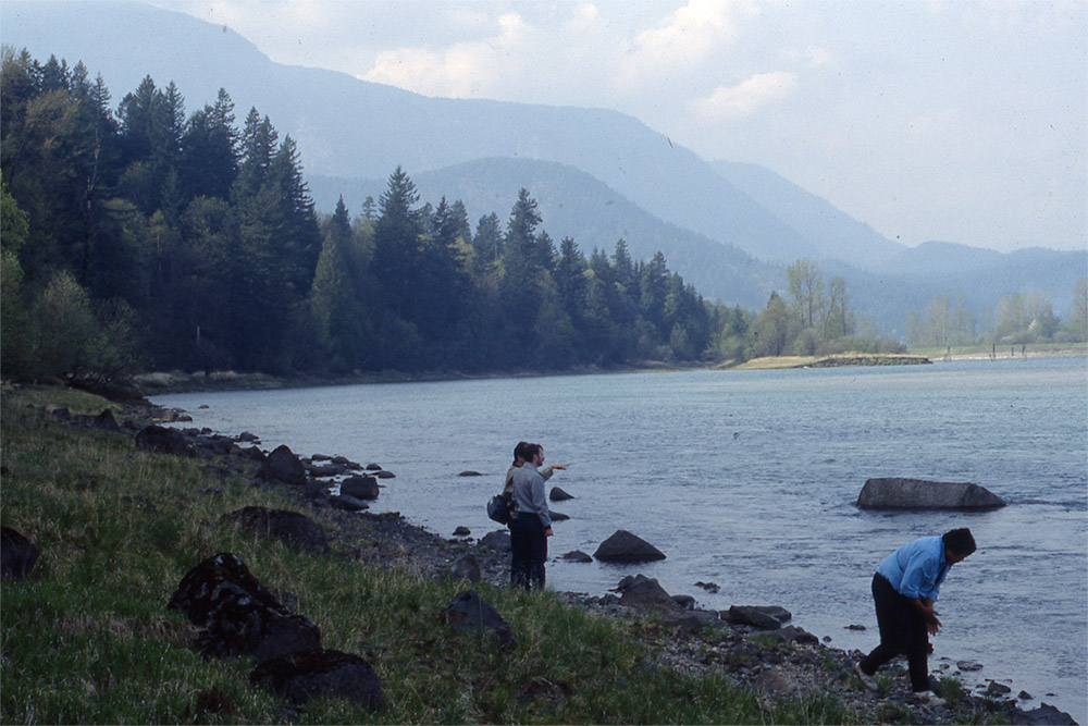 Two people stand on the shoreline and are looking out towards the river. A third person is bent over and looking at the ground.