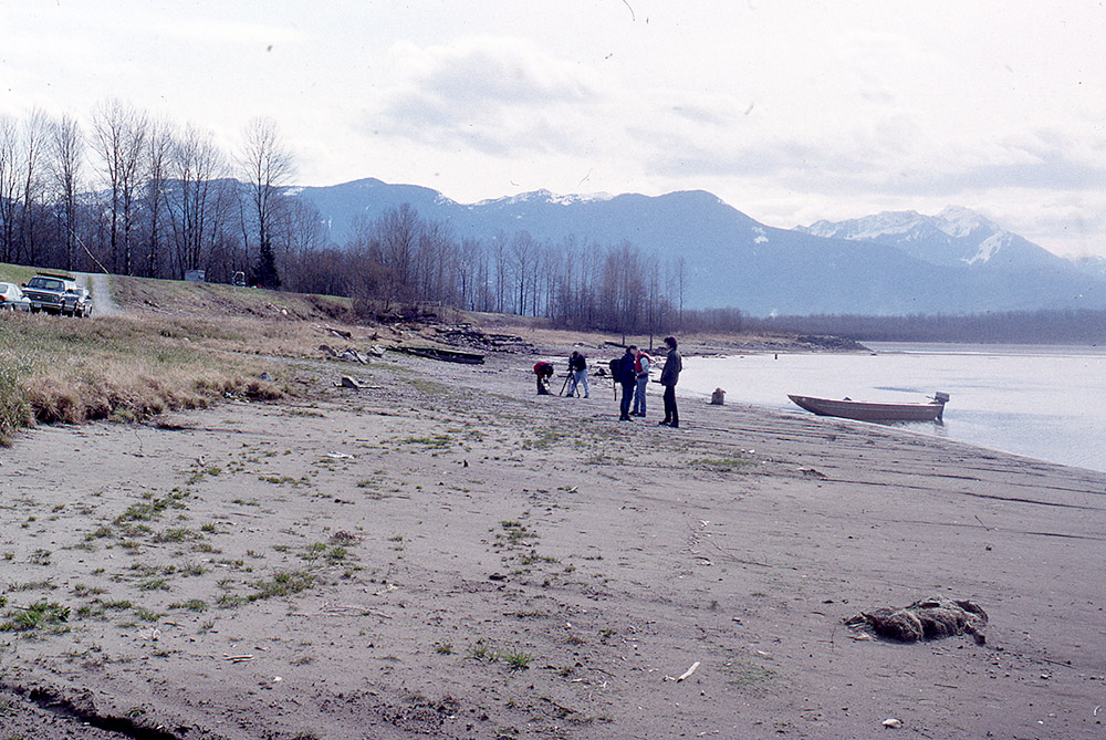 A group of people stand on the shoreline; a small motorboat has been pulled up onto the beach.