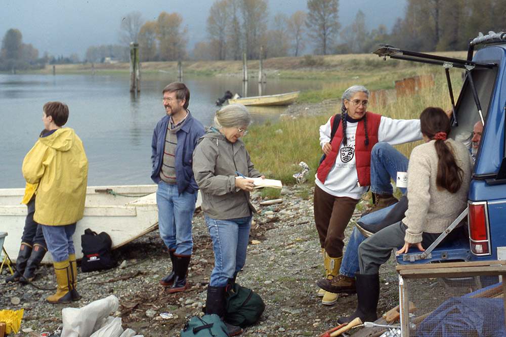 On the shoreline, a group of people have a conversation while they stand by the back of a blue truck.