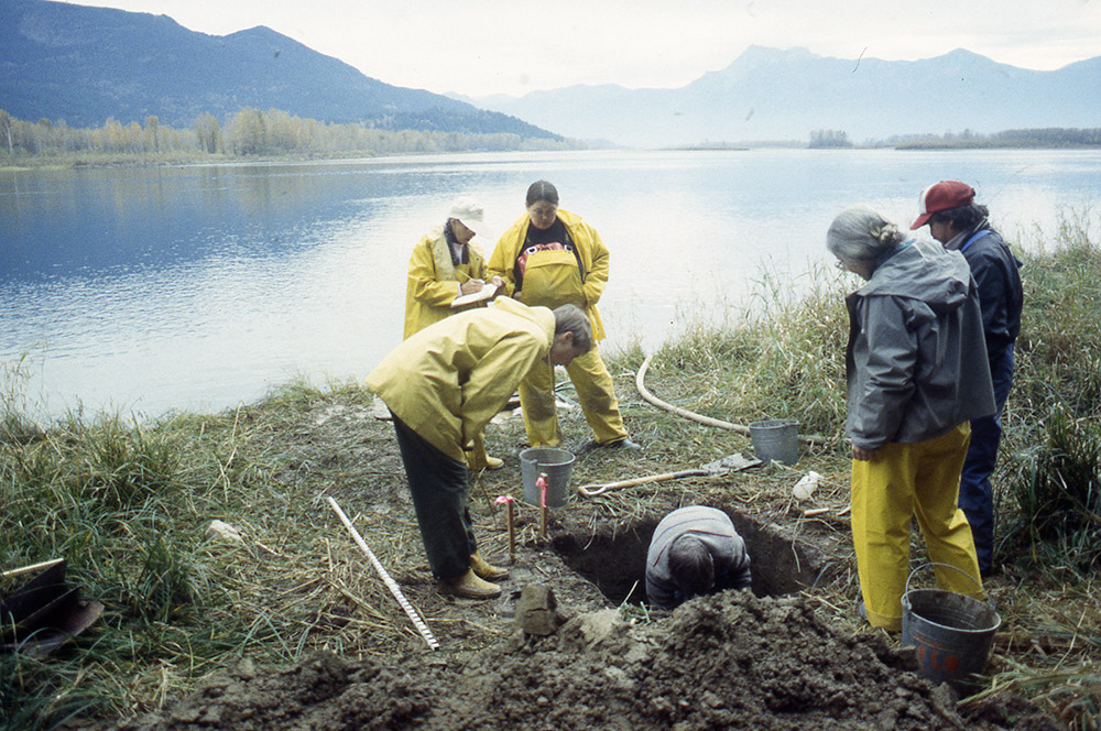 A group of people watch as one person excavates a section of earth on the riverbank.