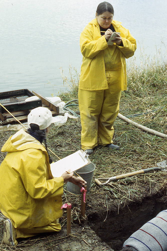 Two archaeologists stand at the edge of an excavation area by the riverbank. They are both taking notes.