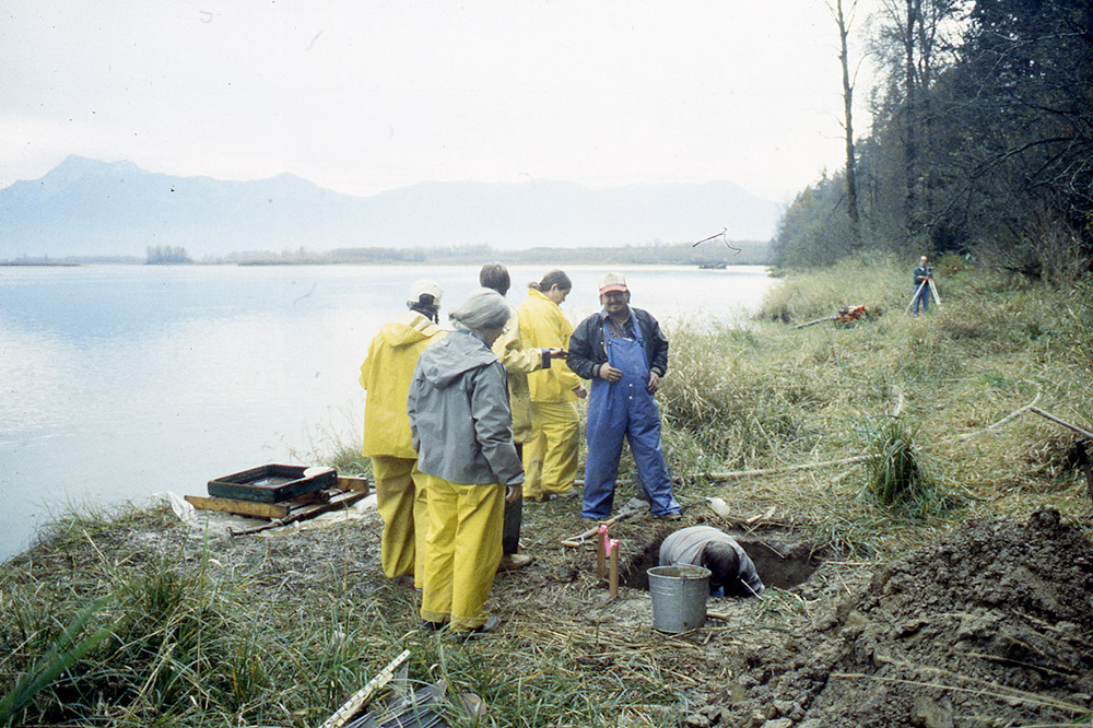 A group of people watch as one person excavates a section of earth on the riverbank.