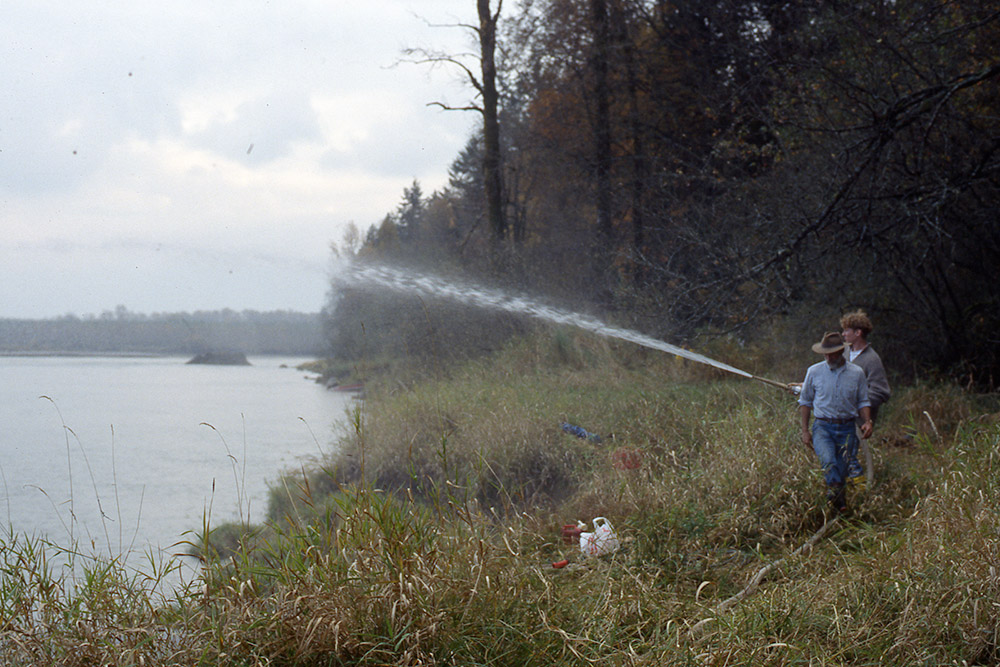 Two people stand on the riverbank. One holds a hose that is spraying a large stream of water into the air towards the river.