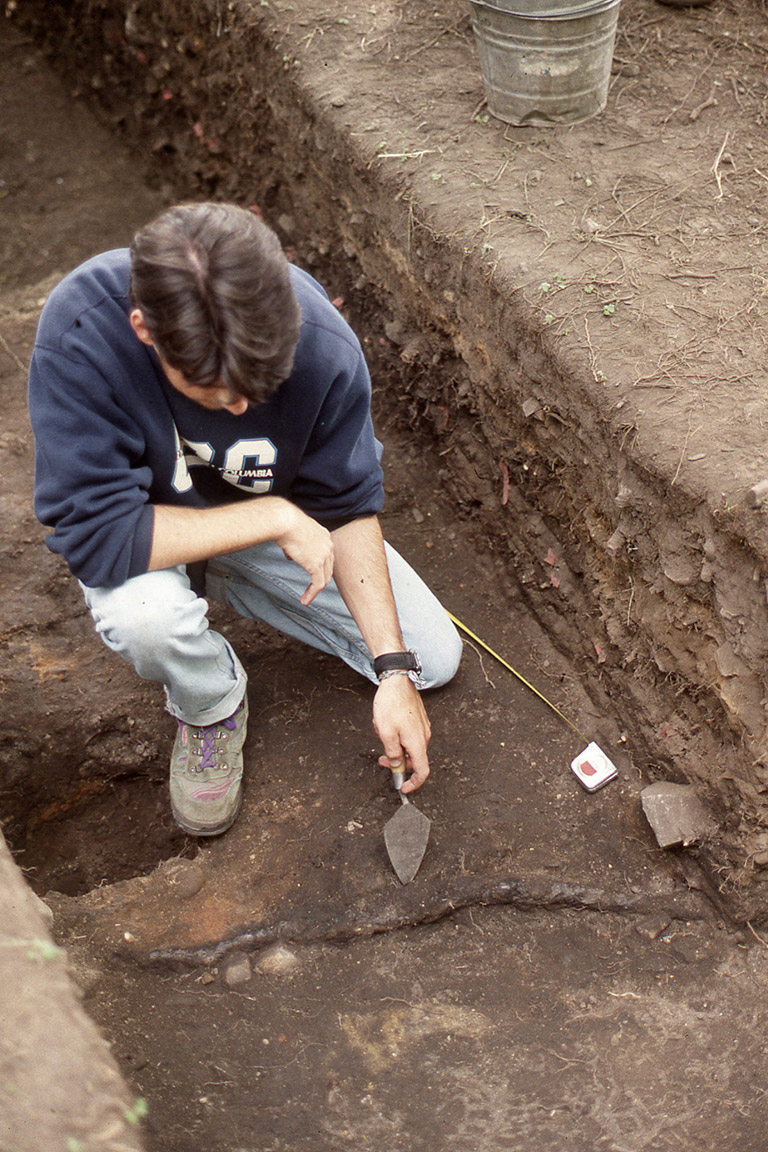 A person examining an archaeological excavation site with a trowel in hand.