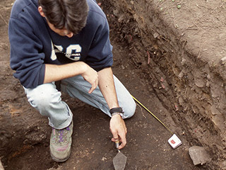 A person examining an archaeological excavation site with a trowel in hand.