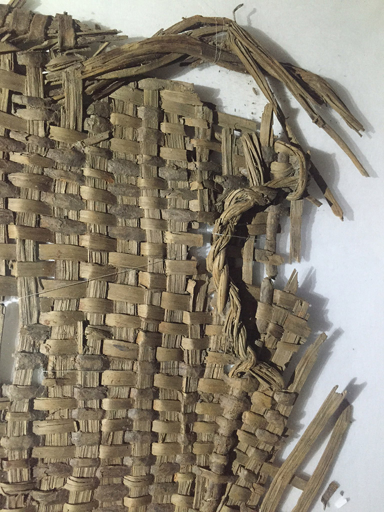 A fragment of  basketry with a woven handle on upper right. The color is light greyish brown.