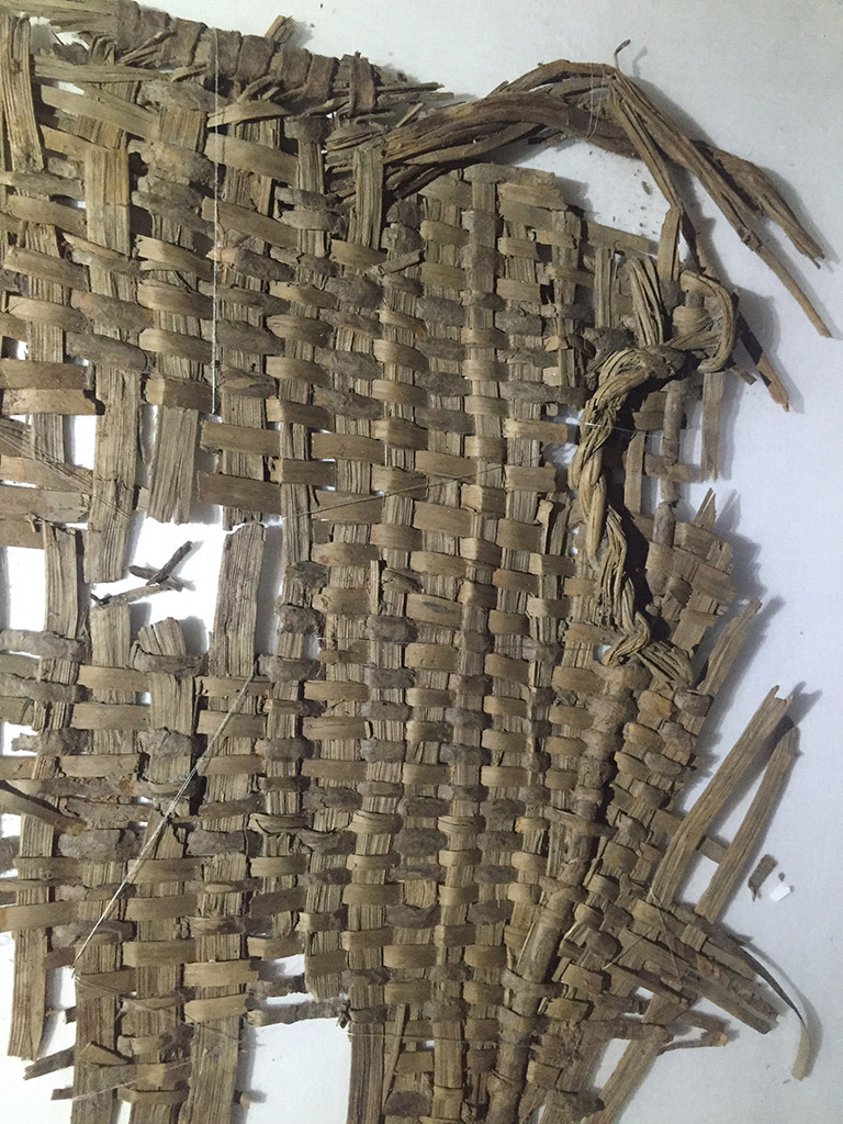 A fragment of woven basketry. The weaving material is light greyish brown.