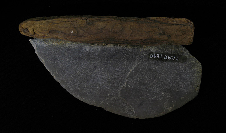 Une pierre gris foncé façonnée en demi-cercle aiguisé, avec un manche de bois qui en est détaché.