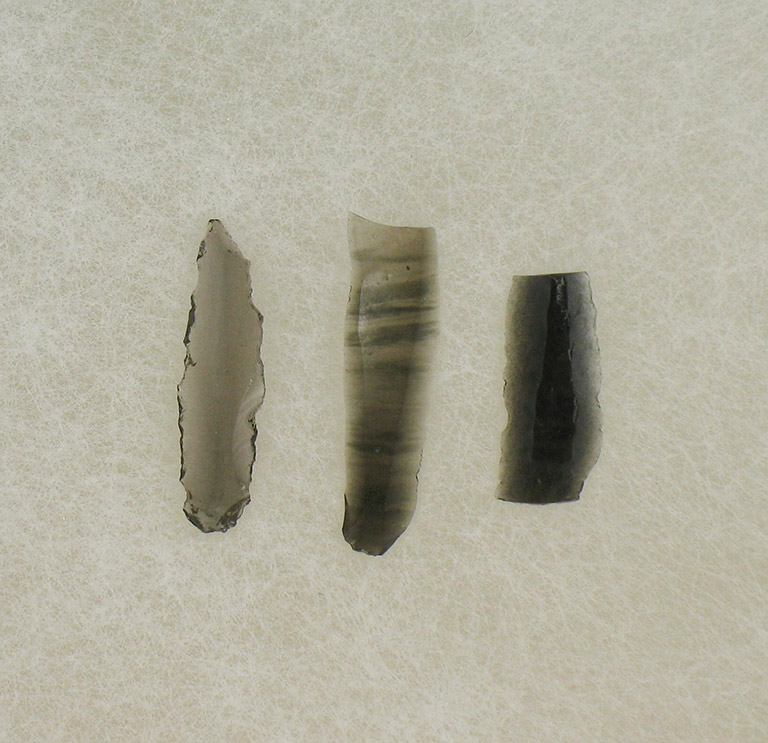 Three thin pieces of translucent rock are shaped into flat blades. They are varying shades of black to grey.