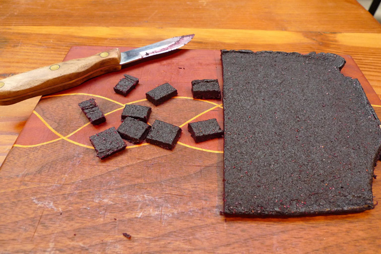 Un gros carré de pâtes de fruits et de plus petits carrés sur une planche à découper. Sur le coin de la planche, il y a un couteau.