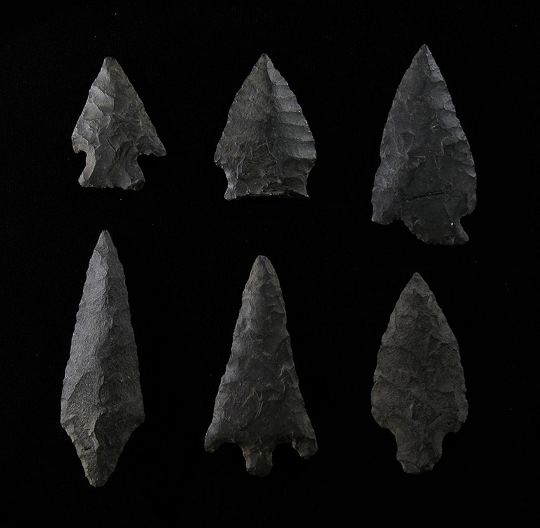Six pointes de flèche de grosseurs différentes faites de pierre gris foncé, posées sur un fond noir.