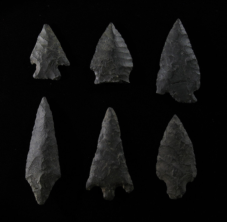 Six arrowheads of different sizes made of dark grey stone on a black background.