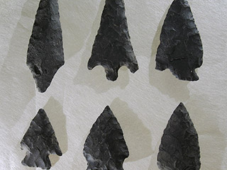 Six pointes de flèche de grosseurs et largeurs différentes faites de pierre foncée, posées sur un fond blanc. 