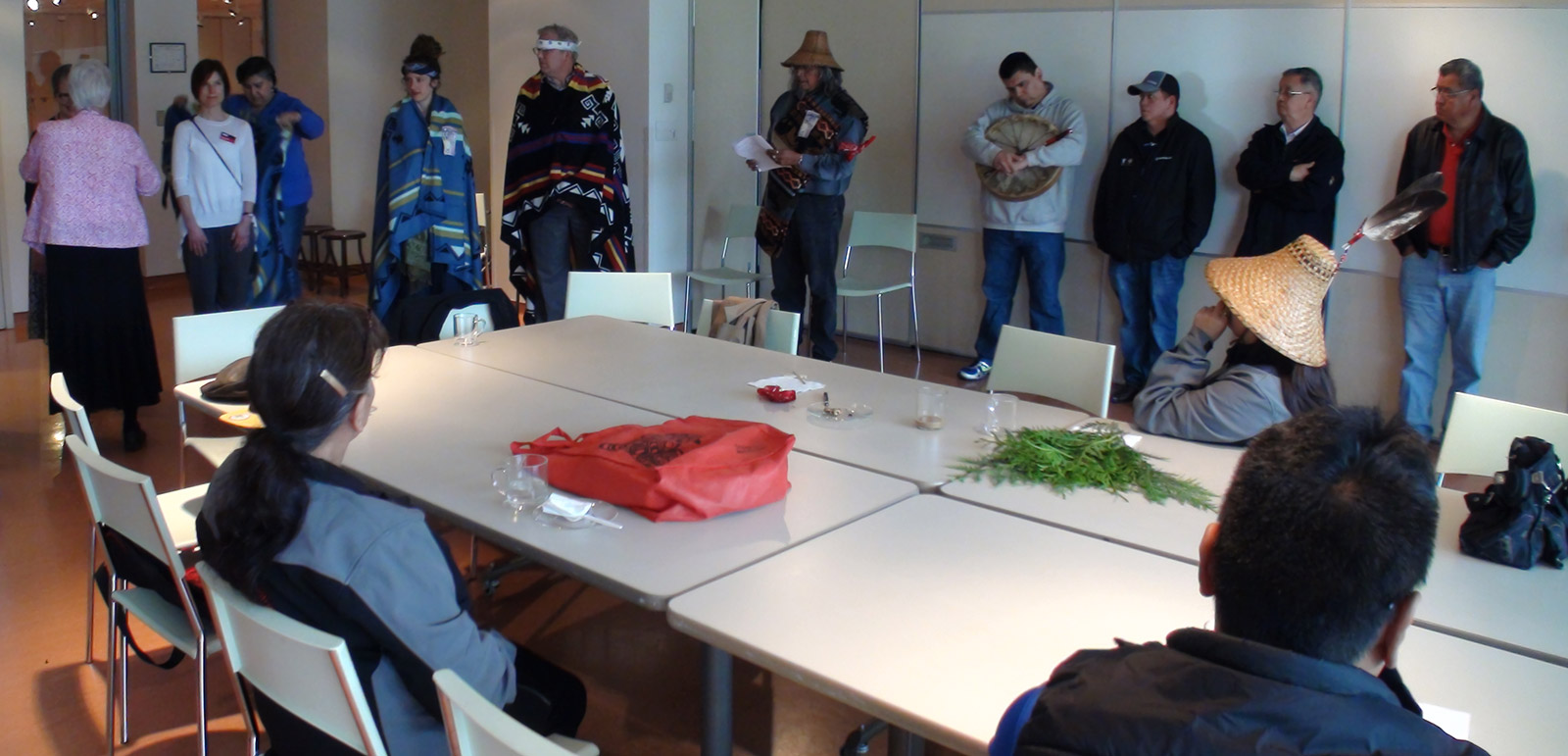 A red bag and some cedar boughs lie on a table. Three people sit at the table, while others stand alongside.