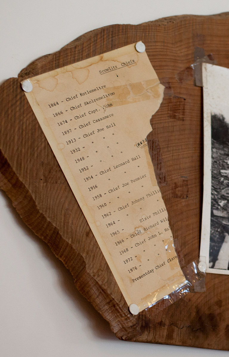 An old ripped piece of paper and a black and white photograph are taped to a piece of wood. The old piece of paper is a list of “Scowlitz Chiefs.” The black and white photograph is of a river bank and forest.