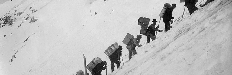 A black and white photograph shows men hiking up a steep snow-covered slope. Each man carries a large pack on his back.