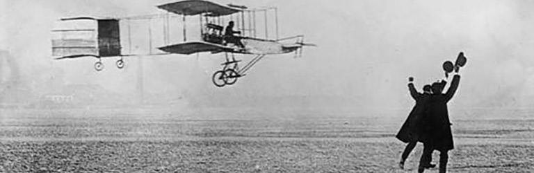 Un biplan vole tout près du sol. À droite, deux hommes ont les bras et le chapeau dans les airs en signe de célébration.