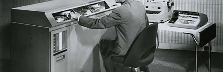 Man sitting in an office with two typewriters and massive square machine with buttons and dials