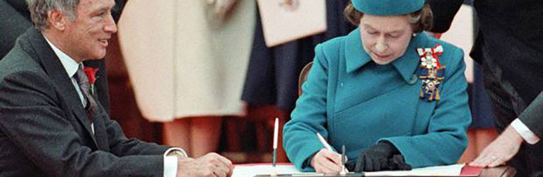 Queen Elizabeth, dressed in a blue hat and coat, signs a document with the Canadian Prime Minister.