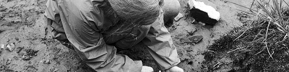 Une photo en noir et blanc d’une femme dans un secteur archéologique. Elle vaporise de l’eau sur les vestiges de vannerie pour les exposer.