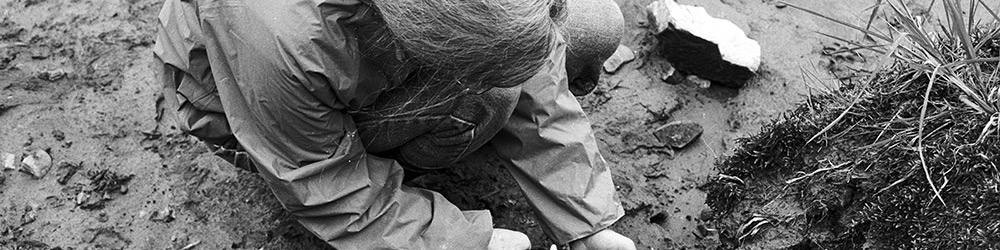 A black and white photograph of a woman in an archaeological area. She is spraying ancient basketry remains with water to expose them. 