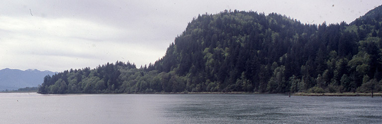 Au premier plan, une rivière aux eaux grisées et sur la rive opposée, une colline basse qui s’élève brusquement pour former une bosse.