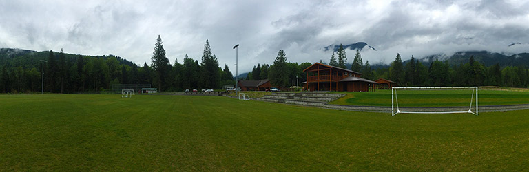 A soccer pitch is in the foreground of the image, with trees and mountains in the background. The sky above is grey and cloudy.