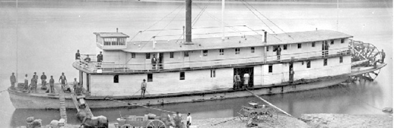 A paddlewheel boat is close to shore. There is a large mountain and forest in the background with a rocky shoreline.