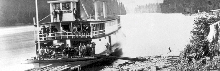 The stern of a paddlewheel boat is pulled close to a rocky shore on the Fraser river, with people on board. There are trees on both sides of the river.