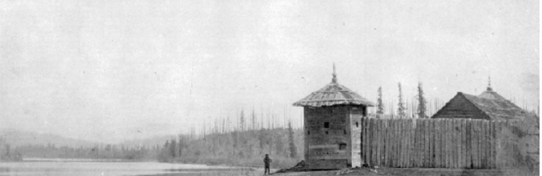  A fort structure is in the mid-ground with a wall of logs in front of it and a person standing outside. There are conifers along the distant shoreline.