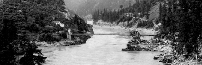 A black and white photograph of a bridge crossing the Fraser River.  The banks of the river are rocky with trees growing up the slopes.
