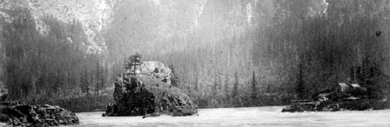 A black and white photograph of a large rock in the middle of the Fraser River with large mountains in the background.