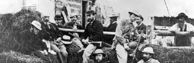 A group of nine men with their guns resting on hay bales sit in front of a house.