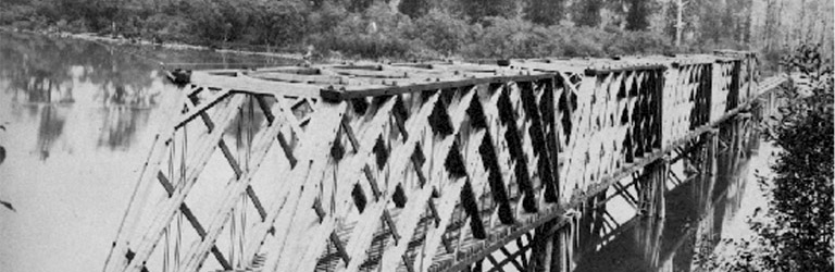 Un ancien pont ferroviaire au-dessus d’une rivière. Il y a des arbres feuillus le long des berges.