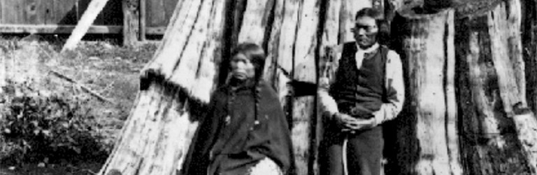 A man and a woman standing in front of a large tree stump. A wooden palisade is in the background. 