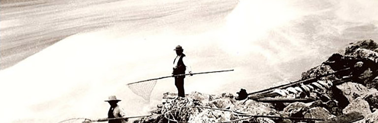 Two people stand on a large rock holding  poles over a whitewater river