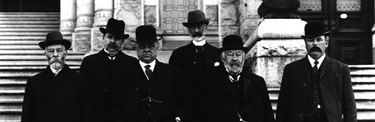 Six hommes en habits et chapeaux debout au bas d’un escalier.